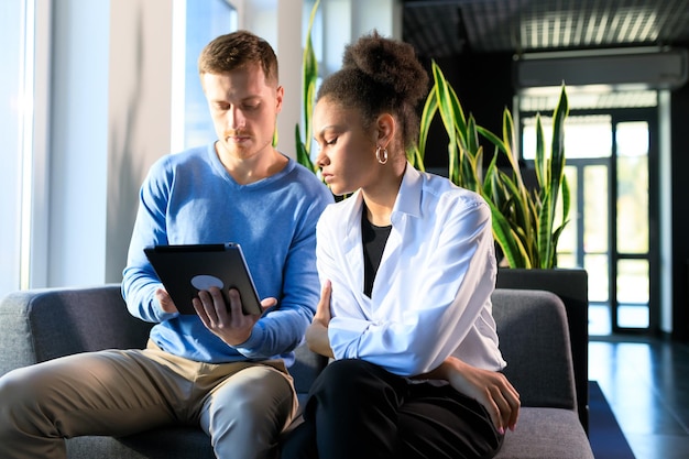 A counselor advising a young adult about different federal job training programs, using a tablet to show the various options available. The setting is a career center with informational posters in the background.